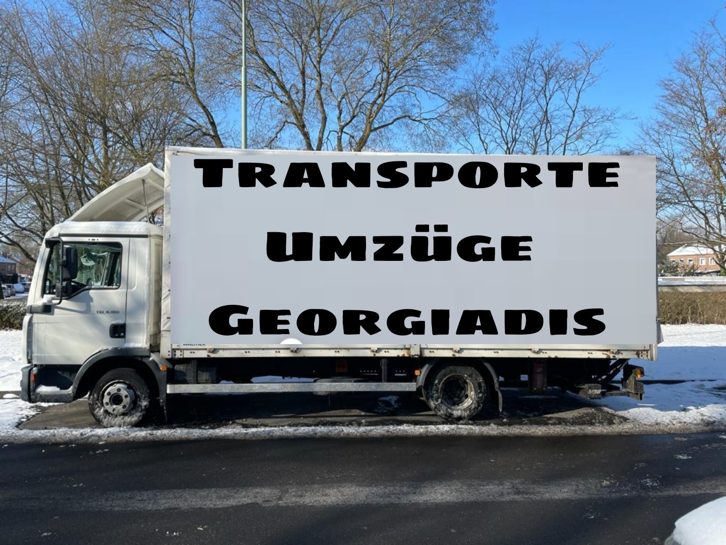 Biała ciężarówka z napisem 'TRANSPORTE UMZÜGE GEORGIADIS' na tle zimowego krajobrazu z drzewami bez liści i pokrytą śniegiem ziemią.