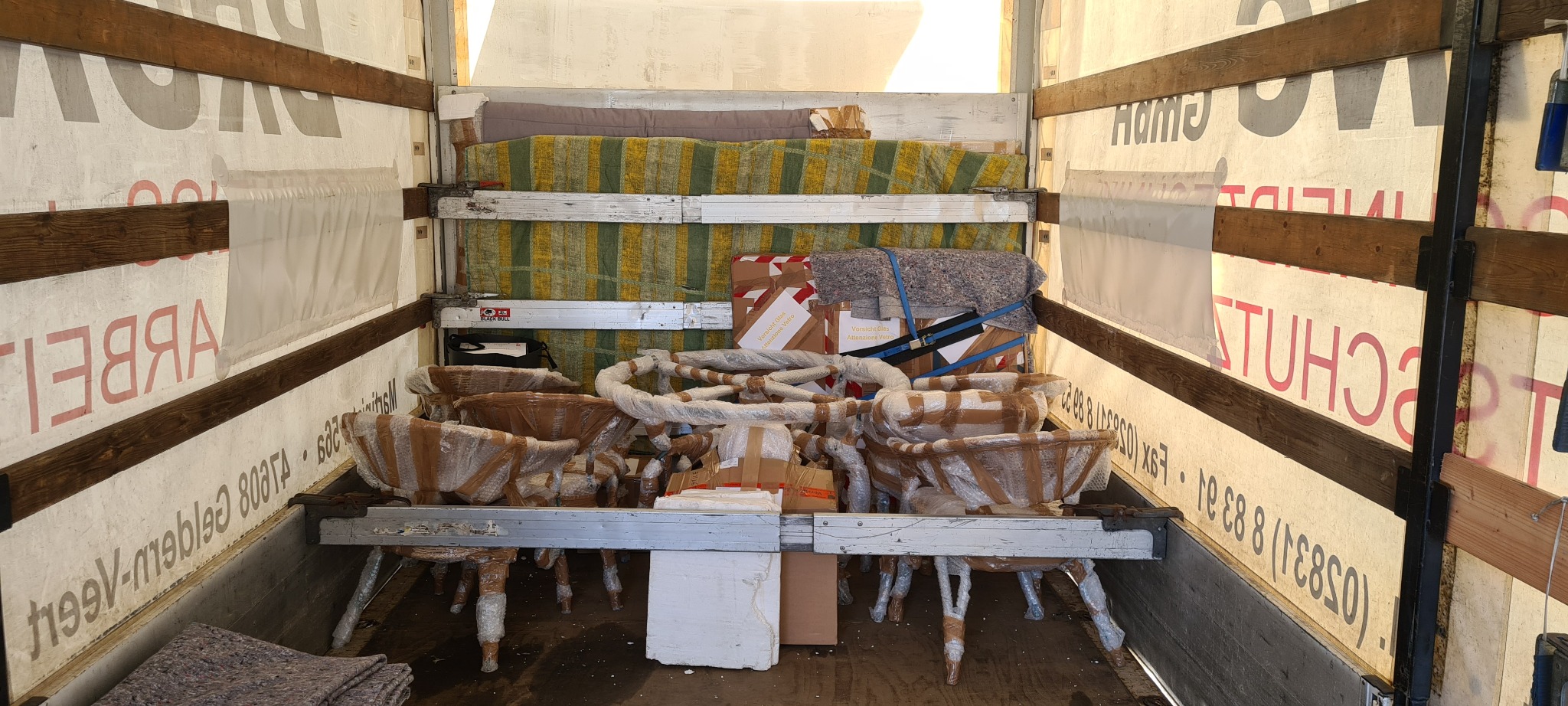 Wnętrze ciężarówki załadowane meblami, w tym krzesłami owiniętymi folią i zabezpieczonymi pasami, kartonami z napisem 'Vorsicht Glas' i kocami ochronnymi, gotowymi do transportu.
