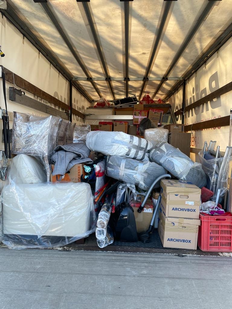 Wnętrze furgonetki załadowane różnorodnymi przedmiotami zabezpieczonymi folią i kartonami, w tym pudełka Archivbox i czerwona plastikowa skrzynka.