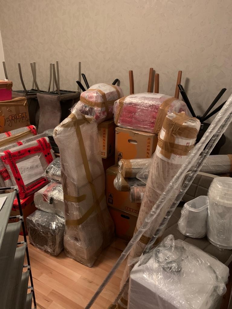 Przygotowane do transportu meble i kartony, owinięte folią i zabezpieczone taśmą, ustawione w pomieszczeniu z widocznymi krzesłami i elementami wyposażenia.