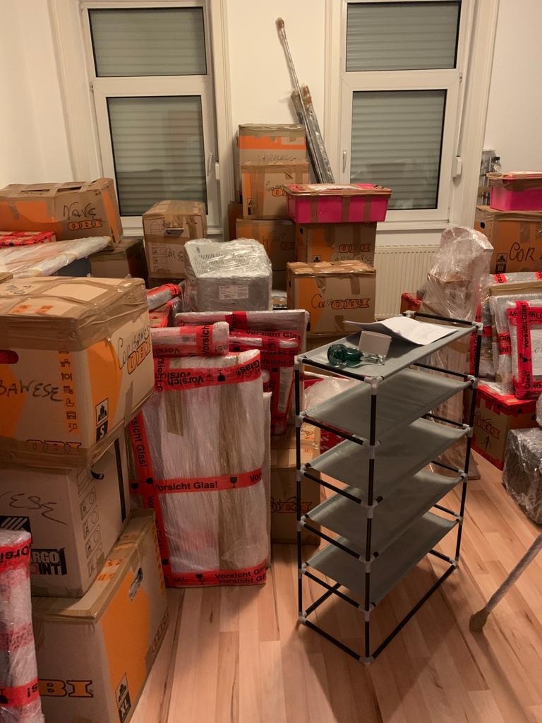 Stos kartonowych pudeł, niektóre z napisem 'Vorsicht Glas!', oraz zdemontowany regał na buty, przygotowane do transportu w mieszkaniu w Köln.