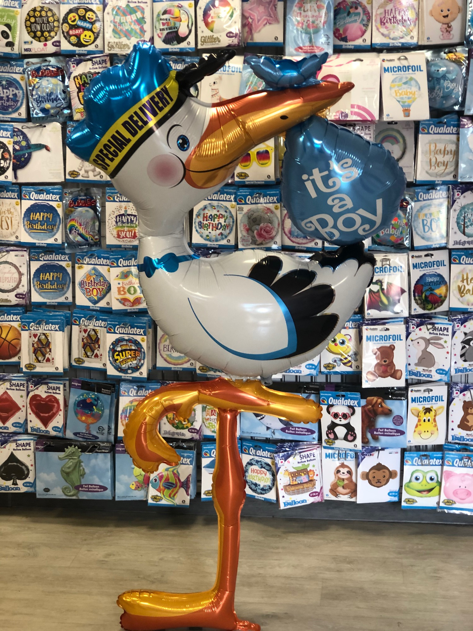 Balon foliowy w kształcie bociana z napisem 'Special Delivery' i niebieskim balonem 'It's a Boy' na tle ściany z różnorodnymi balonami foliowymi.