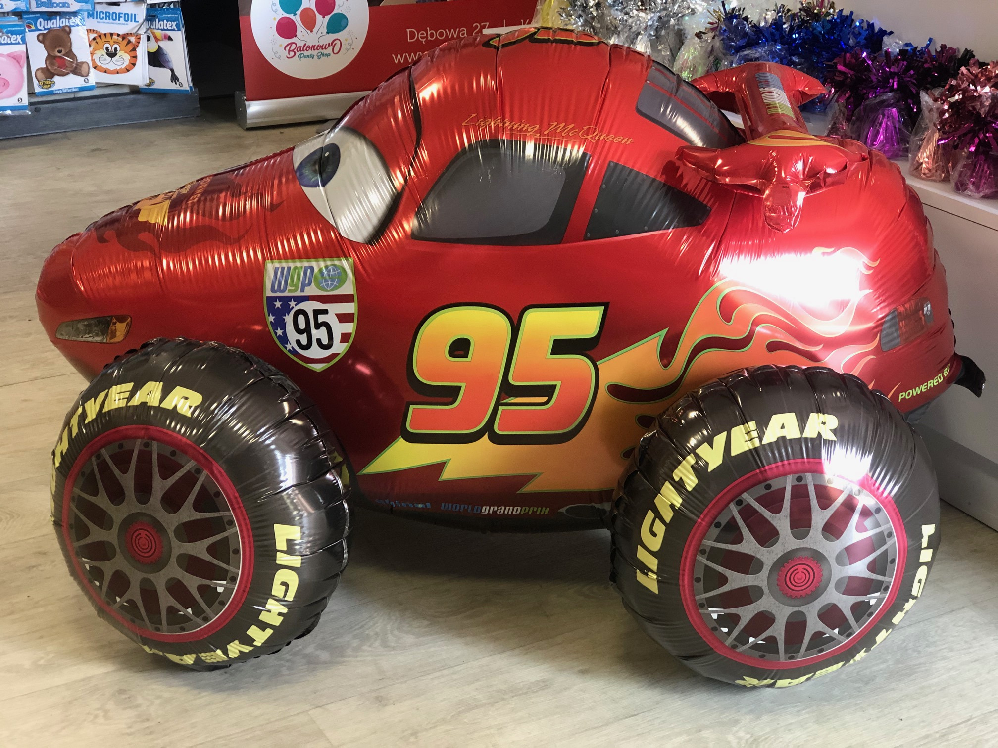 Czerwony balon foliowy w kształcie samochodu Zygzaka McQueen z numerem 95, stojący na podłodze, w tle dekoracje imprezowe i logo sklepu z balonami.