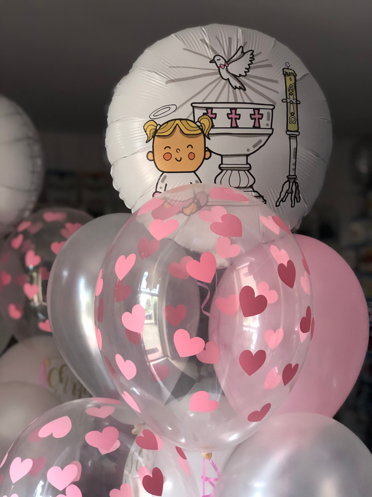 Kompozycja balonowa z motywami religijnymi, w tym balon z rysunkiem dziewczynki z aureolą, gołębia i chrzcielnicy, oraz balony przezroczyste z różowymi serduszkami.