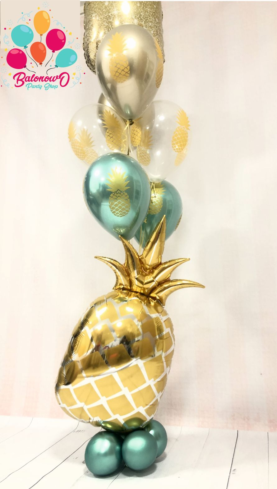 Kompozycja balonowa z dużym balonem w kształcie złotego ananasa i mniejszymi balonami w kolorach złotym, srebrnym i turkusowym z nadrukiem ananasów.