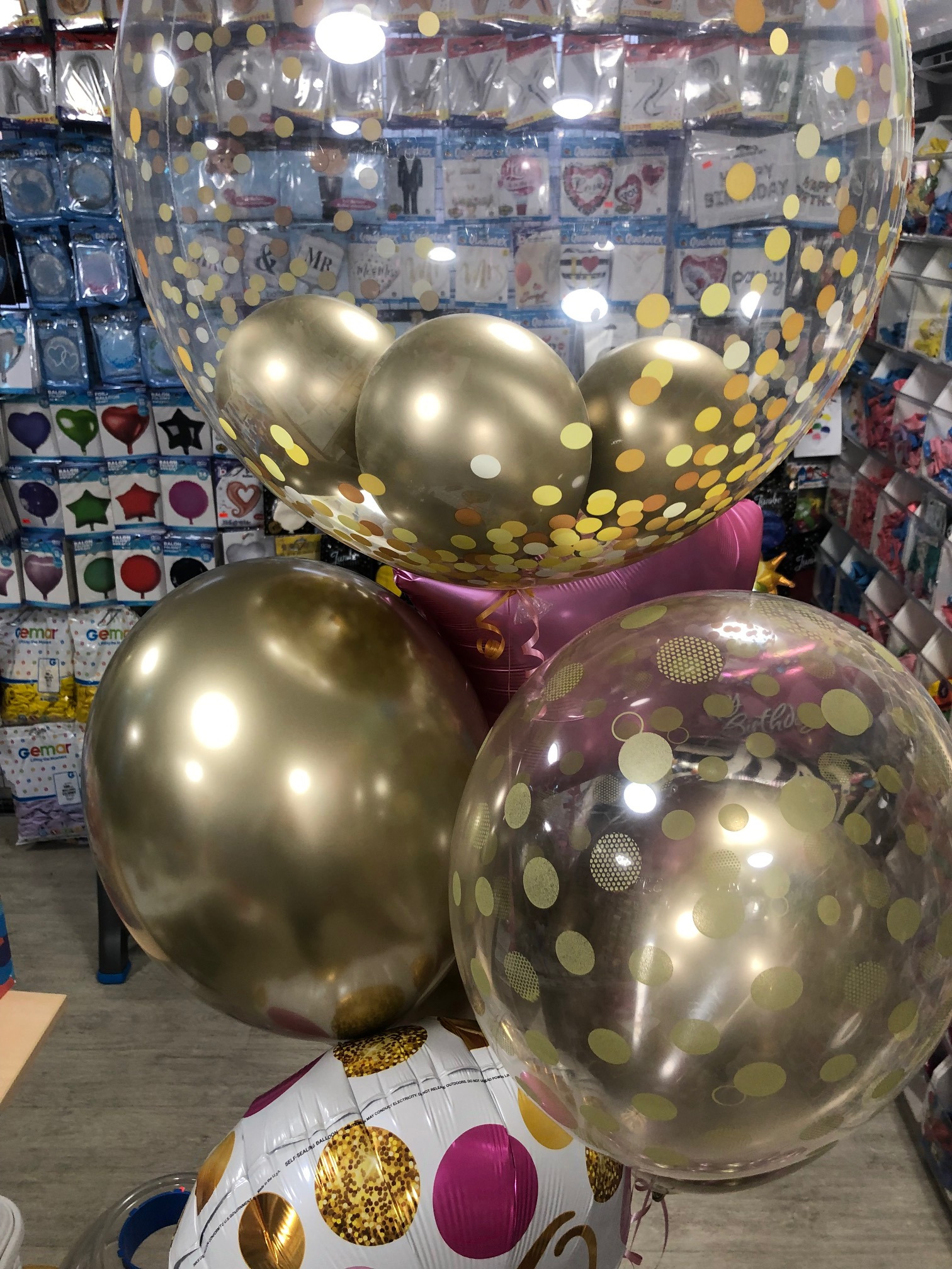 Bukiet balonów: duże złote balony, przezroczysty balon wypełniony złotymi konfetti, oraz biały balon w złote i różowe kropki, na tle sklepowych półek z dekoracjami urodzinowymi.