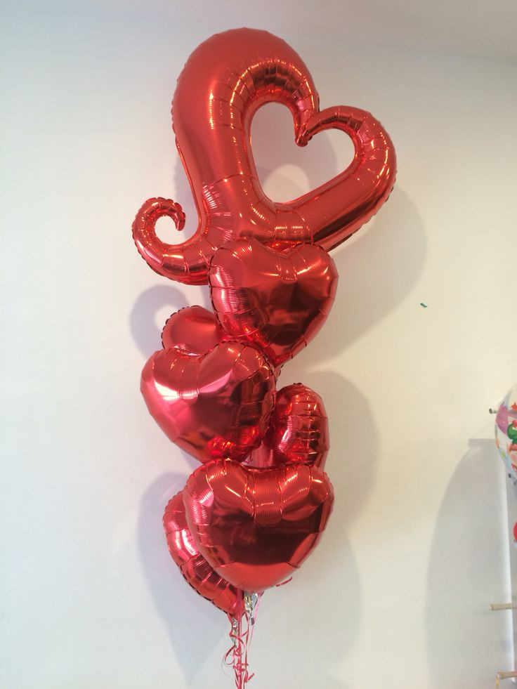 Błyszczący bukiet czerwonych balonów w kształcie serc, z jednym dużym balonem sercem z ozdobnym zawijasem na górze, zawieszony na tle białej ściany.