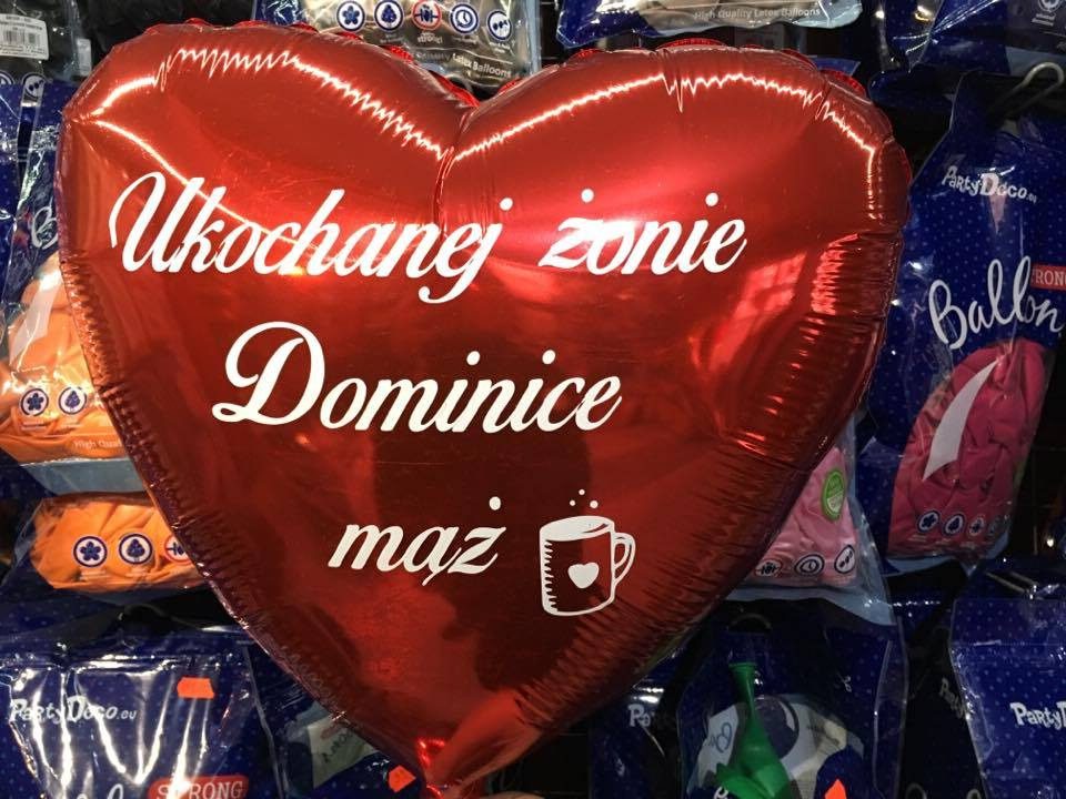 Czerwony balon w kształcie serca z napisem 'Ukochanej żonie Dominice mąż' i rysunkiem kubka z sercem, na tle opakowań balonów.