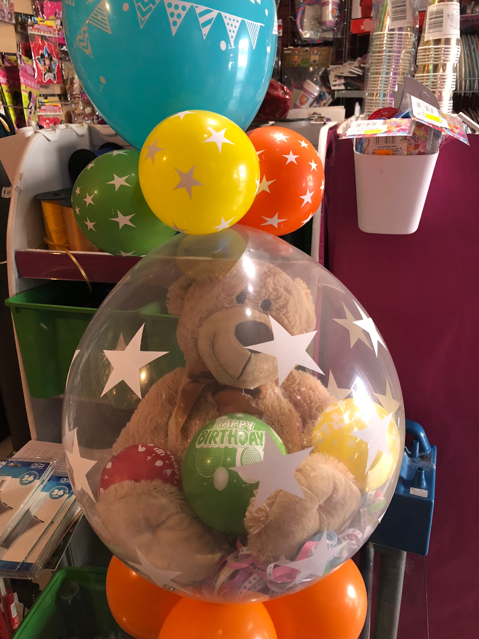 Dekoracja urodzinowa: pluszowy miś w przezroczystym balonie z gwiazdkami, otoczony kolorowymi balonami, w tle wyposażenie sklepu z akcesoriami imprezowymi.
