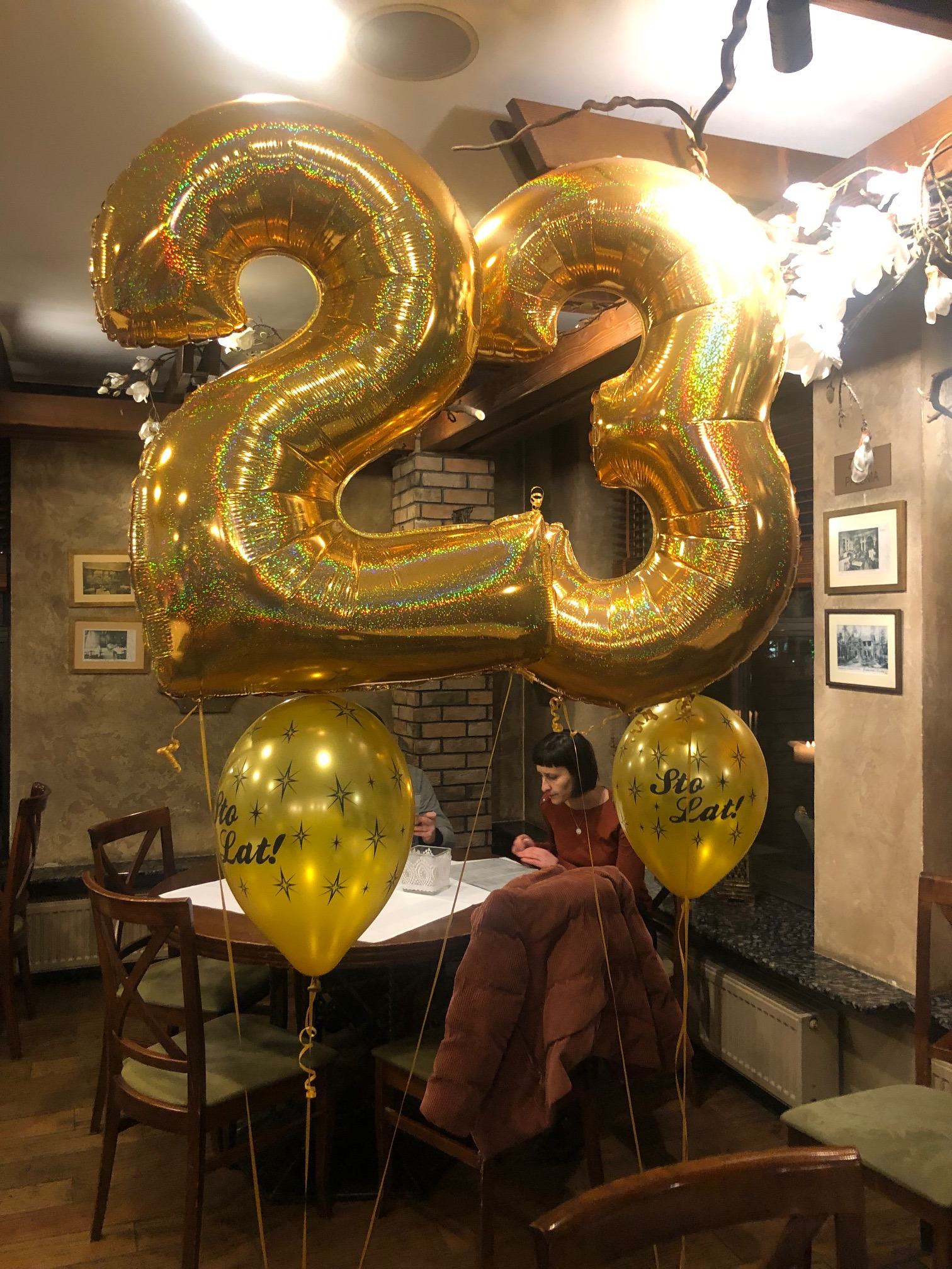 Złote balony w kształcie cyfr 23 zawieszone nad stołem w restauracji, z balonami z napisem 'Sto lat!' i gwiazdkami, w tle siedzące osoby.