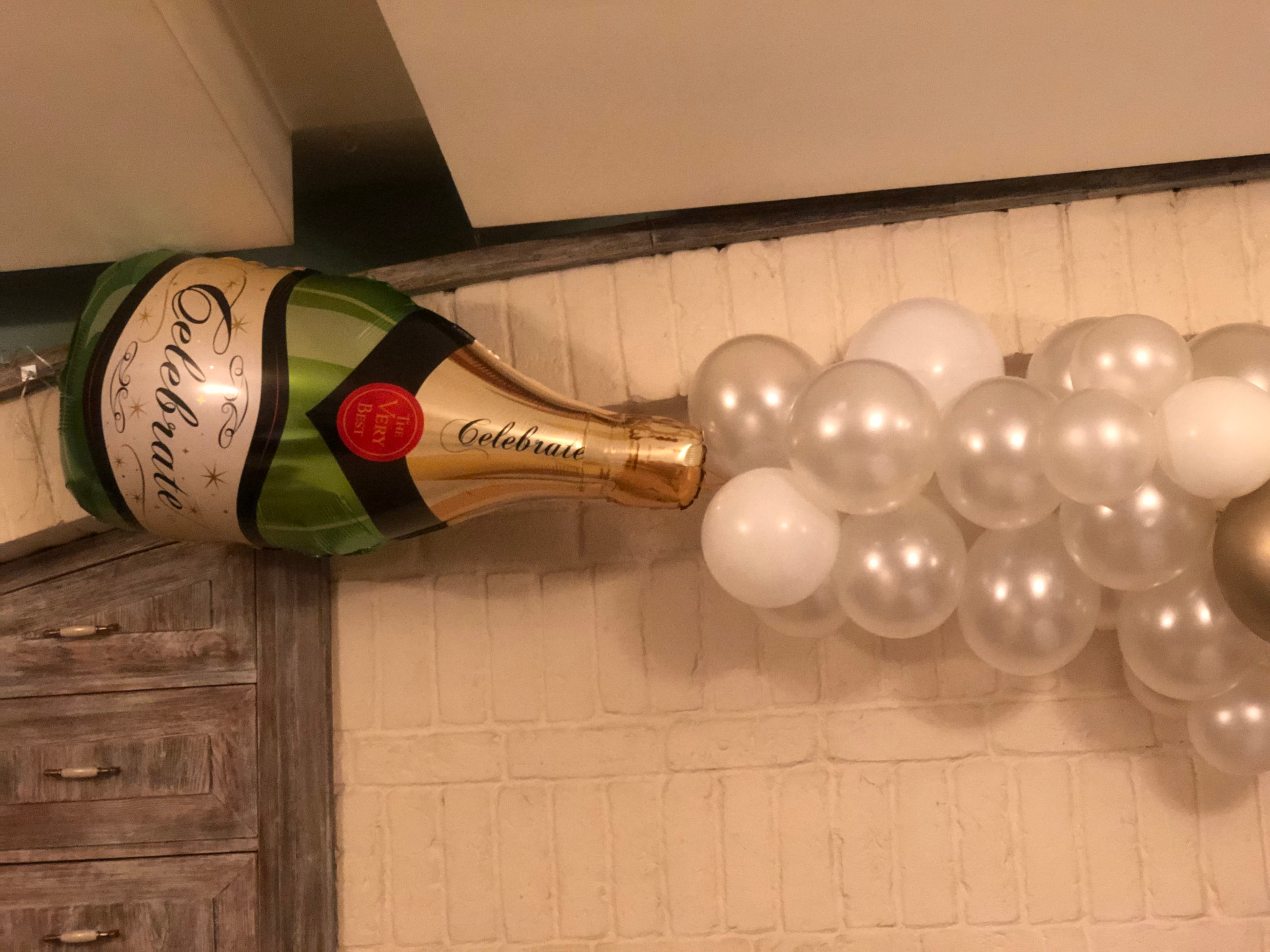 Dekoracja z balonów: foliowy balon w kształcie butelki szampana z napisem Celebrate i białe perłowe balony tworzące efekt wylewającego się trunku na tle białej ceglanej ściany i drewnianej komody.