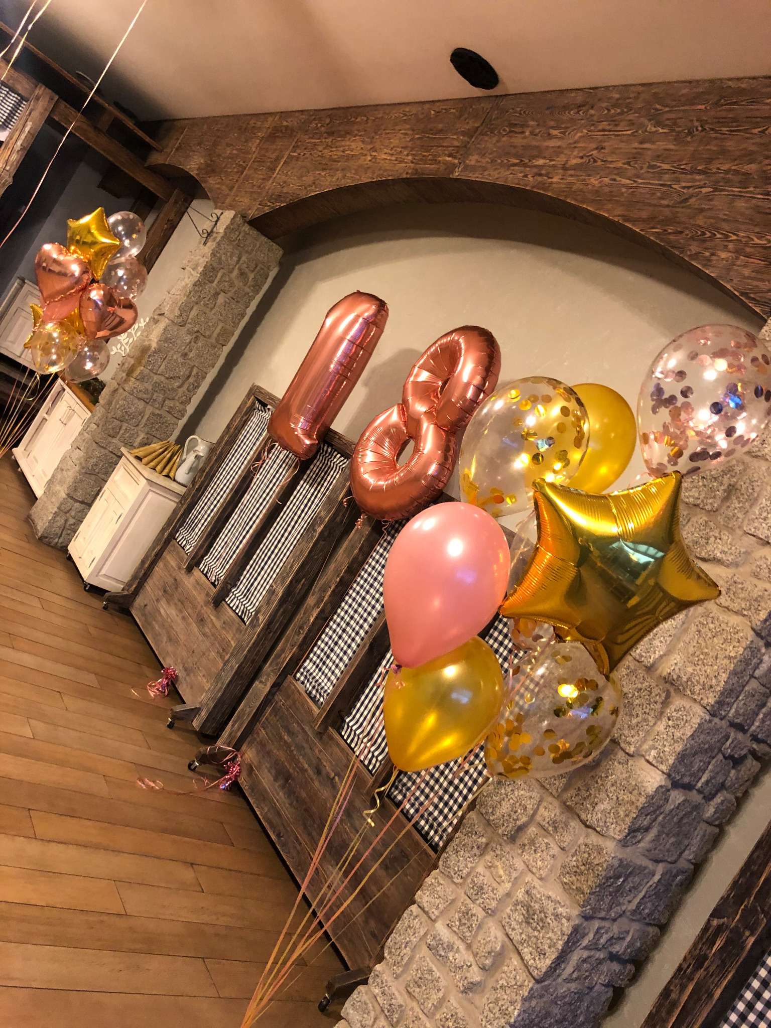 Dekoracja urodzinowa z balonów w kształcie cyfr 1 i 8, złotych gwiazd i przezroczystych kul z konfetti, na tle rustykalnej ściany z kamienia i drewnianej zabudowy z tkaniną w kratę.