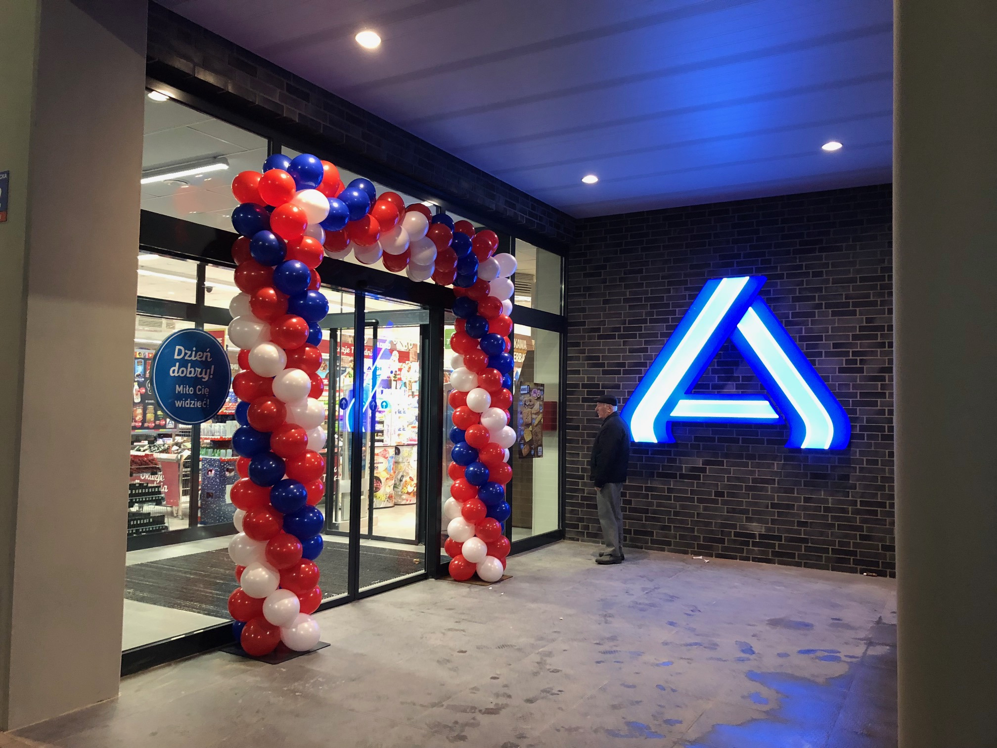 Otwarcie sklepu z dekoracją balonową w kolorach czerwonym, białym i niebieskim, neonowe logo na ceglanej ścianie, klient w tle.