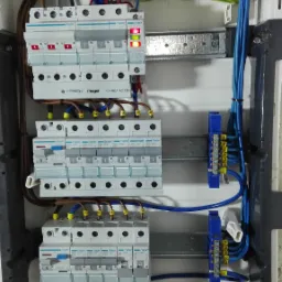 Rozdzielnica elektryczna z zainstalowanymi bezpiecznikami Hager, widoczne okablowanie w kolorach niebieskim, zielono-żółtym i brązowym, jeden z bezpieczników z diodą sygnalizacyjną w kolorze...