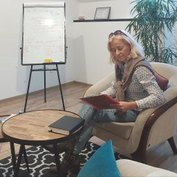 Iwona Wróbel Terapia uzależnień - Kobieta w fotelu czyta dokument w czerwonej teczce, w tle tablica flipchart z notatkami dotyczącymi asertywności i oczekiwań.