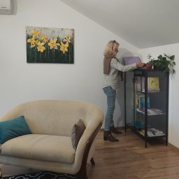 Iwona Wróbel Terapia uzależnień - Kobieta w jasnym swetrze i dżinsach stoi w jasnym pokoju, obok beżowej kanapy z turkusową poduszką i czarnego regału z książkami i roślinami doniczkowymi, na ścianie obraz z żółtymi kwiatami.