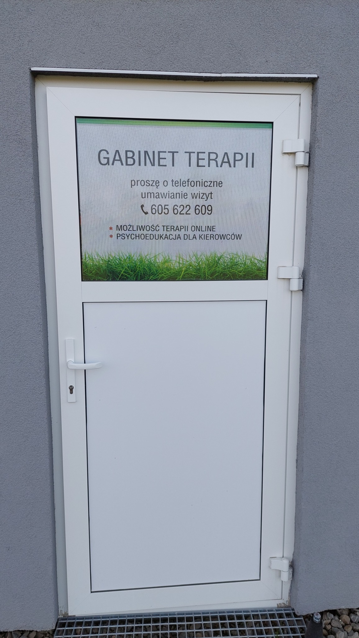Białe drzwi wejściowe do gabinetu z naklejoną informacją o możliwości umówienia wizyty telefonicznie oraz o terapii online i psychoedukacji dla kierowców.