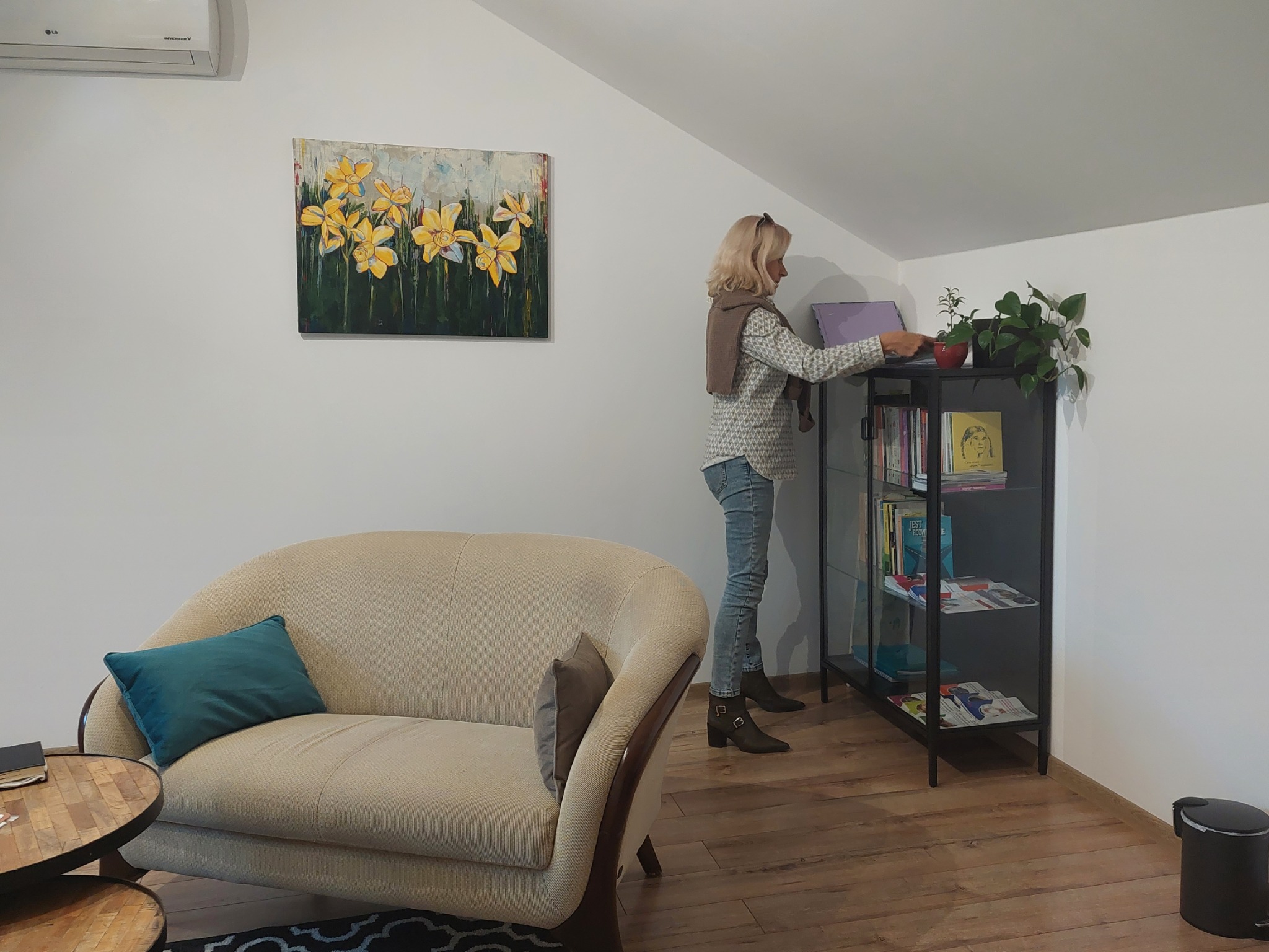 Kobieta w jasnym swetrze i dżinsach stoi w jasnym pokoju, obok beżowej kanapy z turkusową poduszką i czarnego regału z książkami i roślinami doniczkowymi, na ścianie obraz z żółtymi kwiatami.