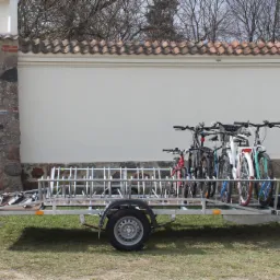 Przyczepa rowerowa z zamontowanymi rowerami różnych kolorów, zaparkowana na trawie przed murem z elementami kamiennymi i ceglaną dachówką.