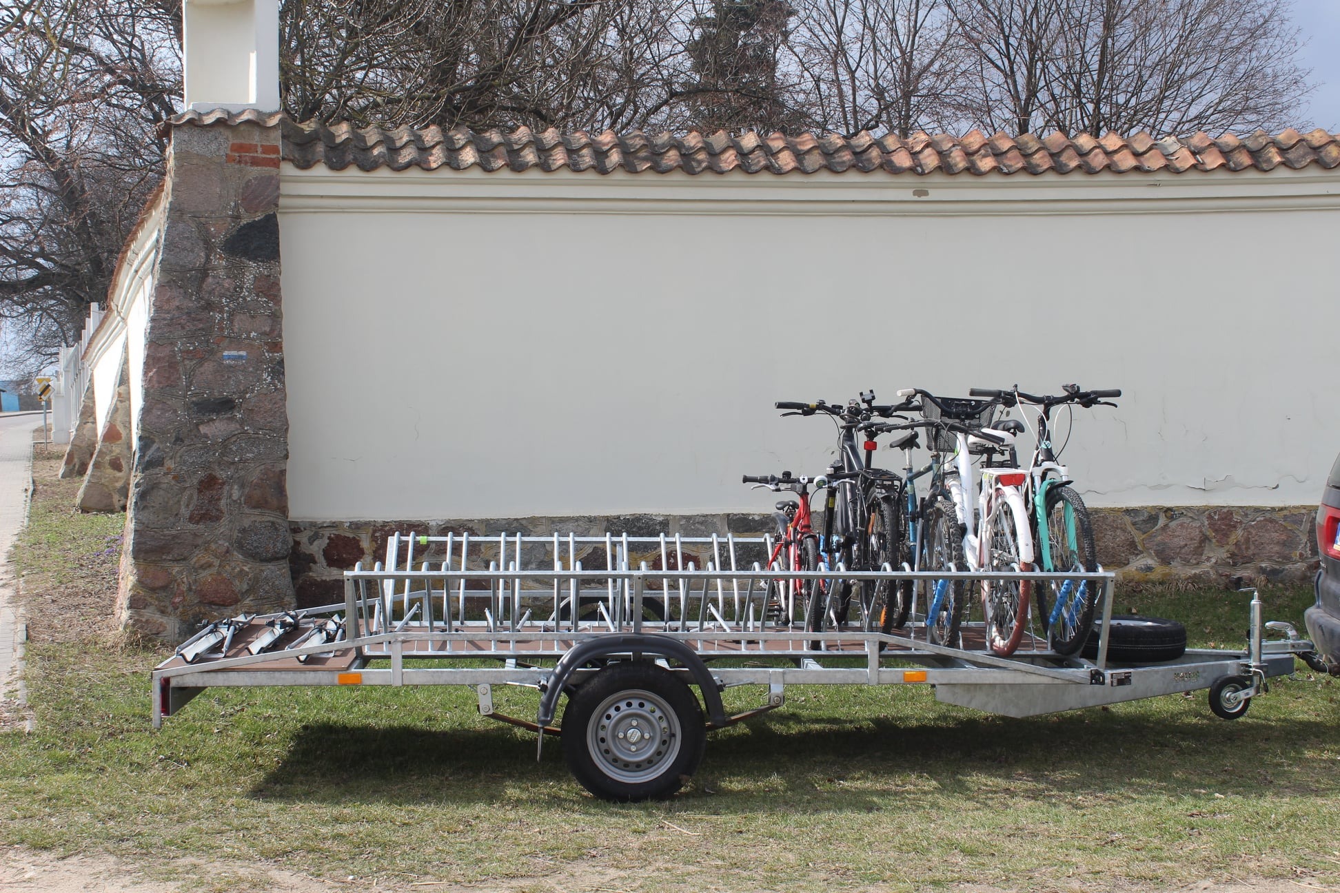 Przyczepa rowerowa z zamontowanymi rowerami różnych kolorów, zaparkowana na trawie przed murem z elementami kamiennymi i ceglaną dachówką.