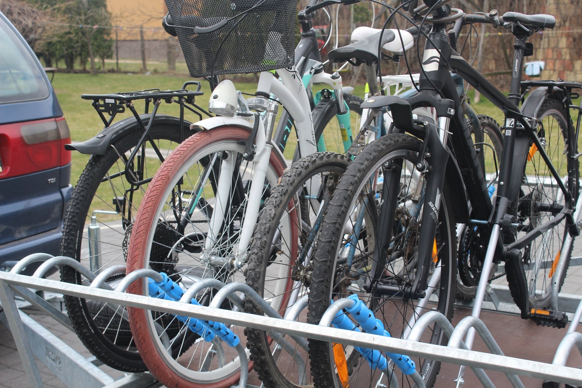 Rząd rowerów różnych typów zaparkowanych w metalowym stojaku z niebieskimi zabezpieczeniami, obok zaparkowany niebieski samochód.