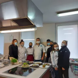 Grupa osób w maskach, ubranych w stroje kucharskie, prezentuje talerze z gotowymi daniami w profesjonalnej kuchni z metalowym okapem.