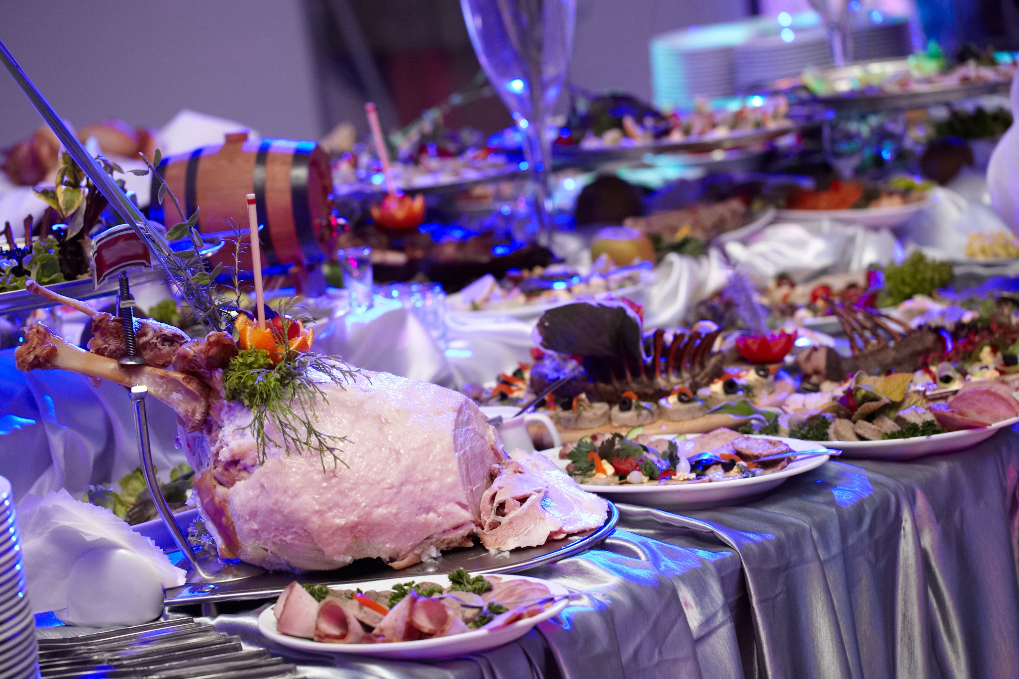 Uroczysty stół szwedzki z pieczonym udźcem dekorowanym ziołami i pomarańczą, w tle tace z przekąskami i mała beczka.