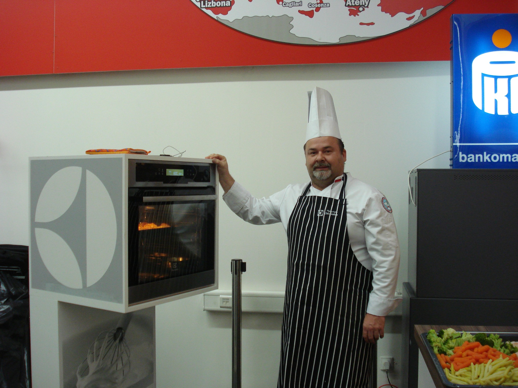 Kucharz w stroju roboczym, z piekarnikiem Electrolux, prezentuje przygotowywanie potrawy w Krakowie. W tle mapa Europy i logo bankomatu.