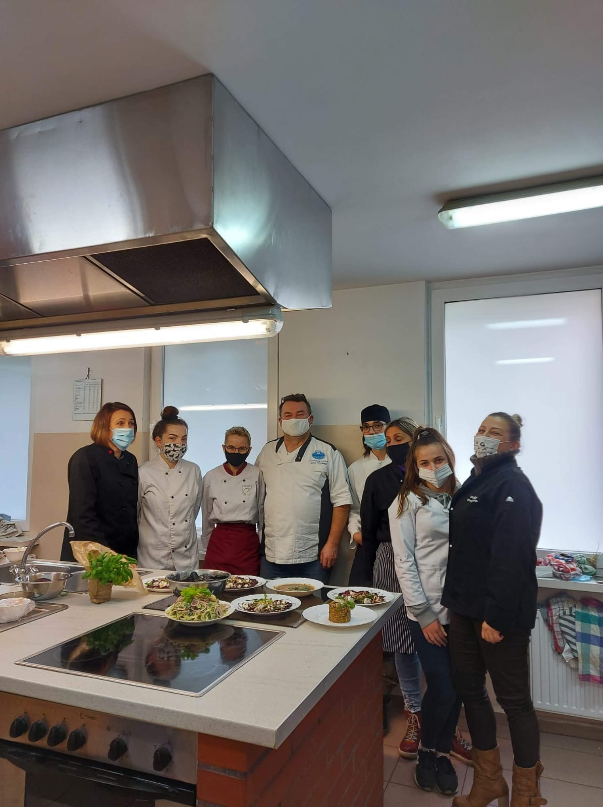 Grupa osób w maskach, ubranych w stroje kucharskie, prezentuje talerze z gotowymi daniami w profesjonalnej kuchni z metalowym okapem.