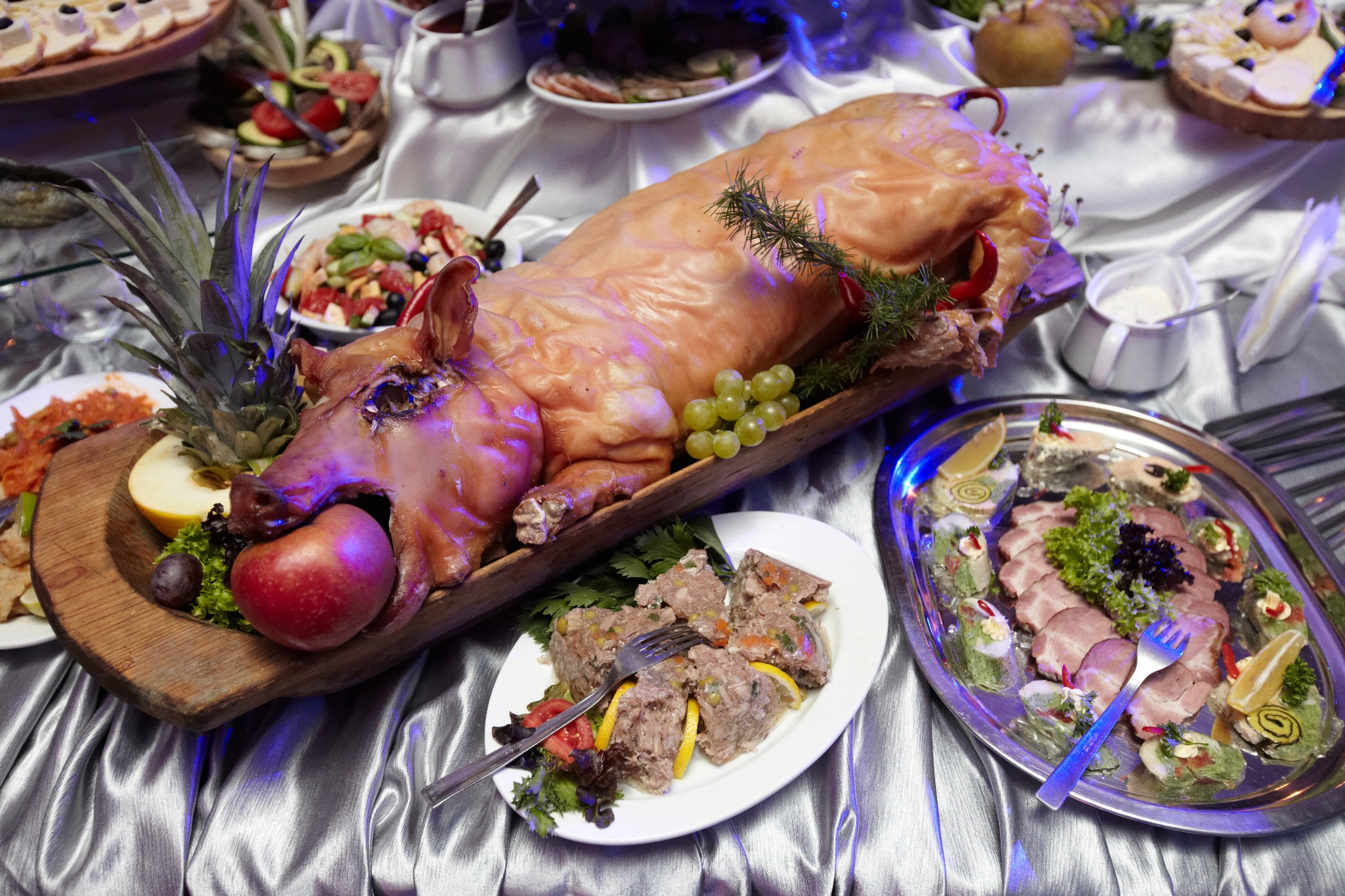 Pieczone prosię z jabłkiem w pysku, dekorowane ananasem i winogronami, otoczone półmiskami z przekąskami na błyszczącym obrusie.