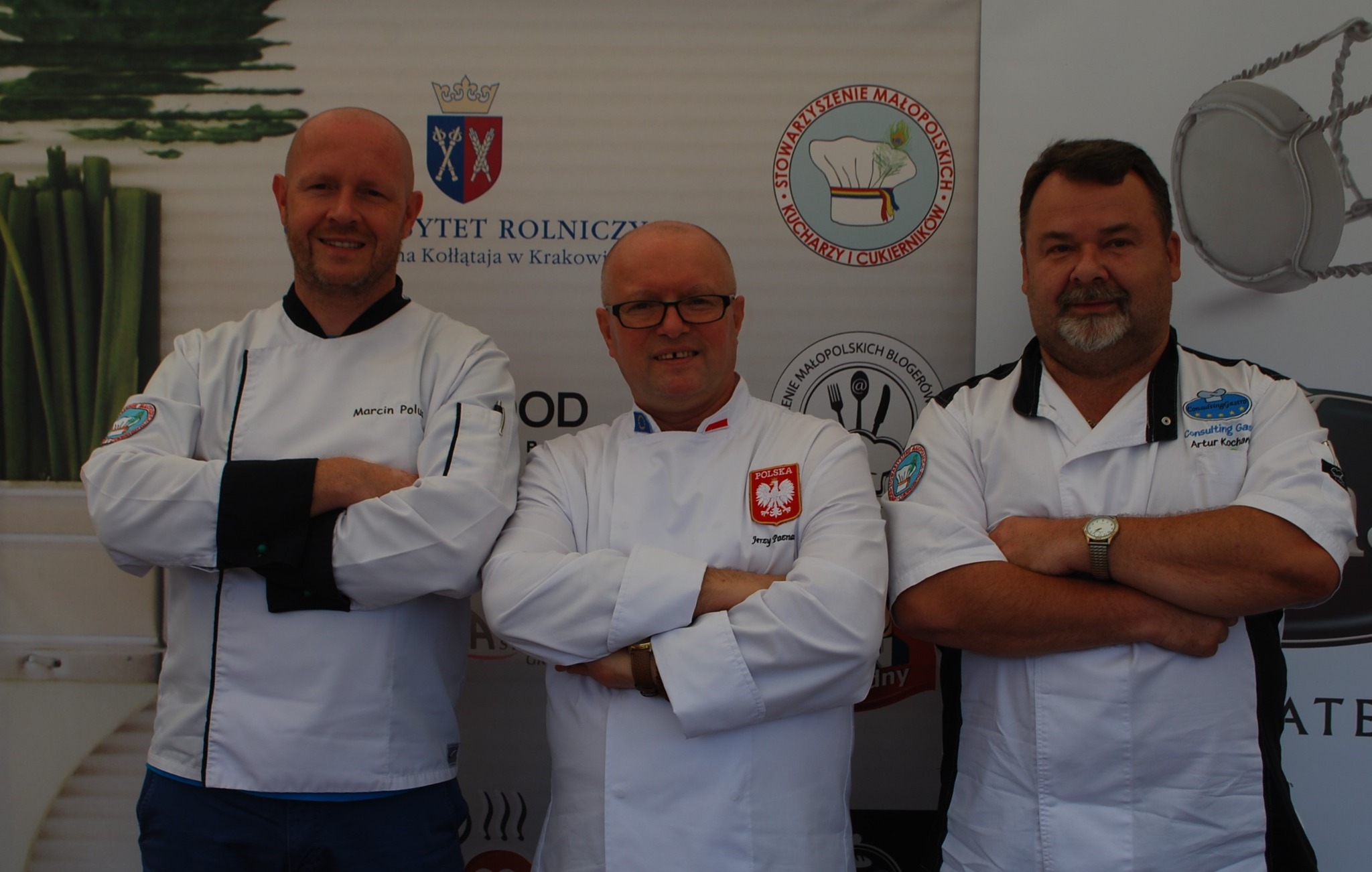 Trzech mężczyzn w strojach kucharzy pozuje na tle baneru z logotypami organizacji kulinarnych, w tym Stowarzyszenia Małopolskich Kucharzy i Cukierników i Instytutu Rolniczego.