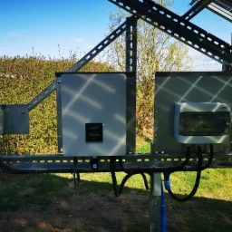 Instalacja fotowoltaiczna: widok na inwerter FoxESS zamontowany na stalowej konstrukcji, obok skrzynka z bezpiecznikami i licznik energii, w tle zielona trawa i krzewy, błękitne niebo.