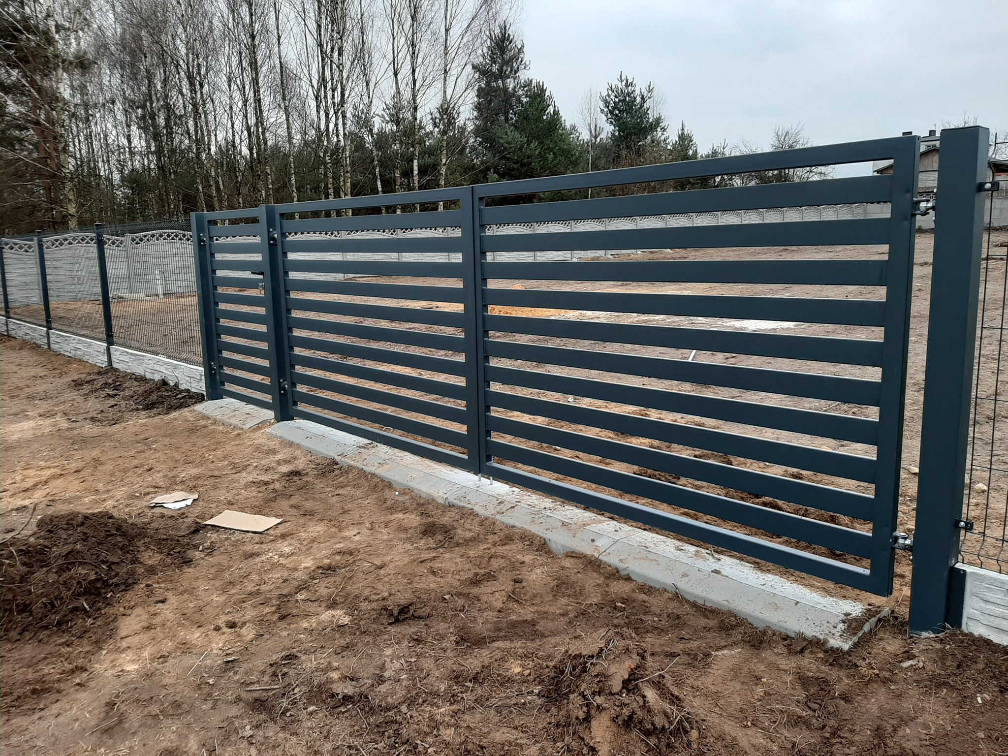 Nowoczesne, poziome, szare przęsła ogrodzeniowe z metalu, osadzone na betonowej podmurówce, widoczne na tle siatki ogrodzeniowej i drzew bezlistnych.