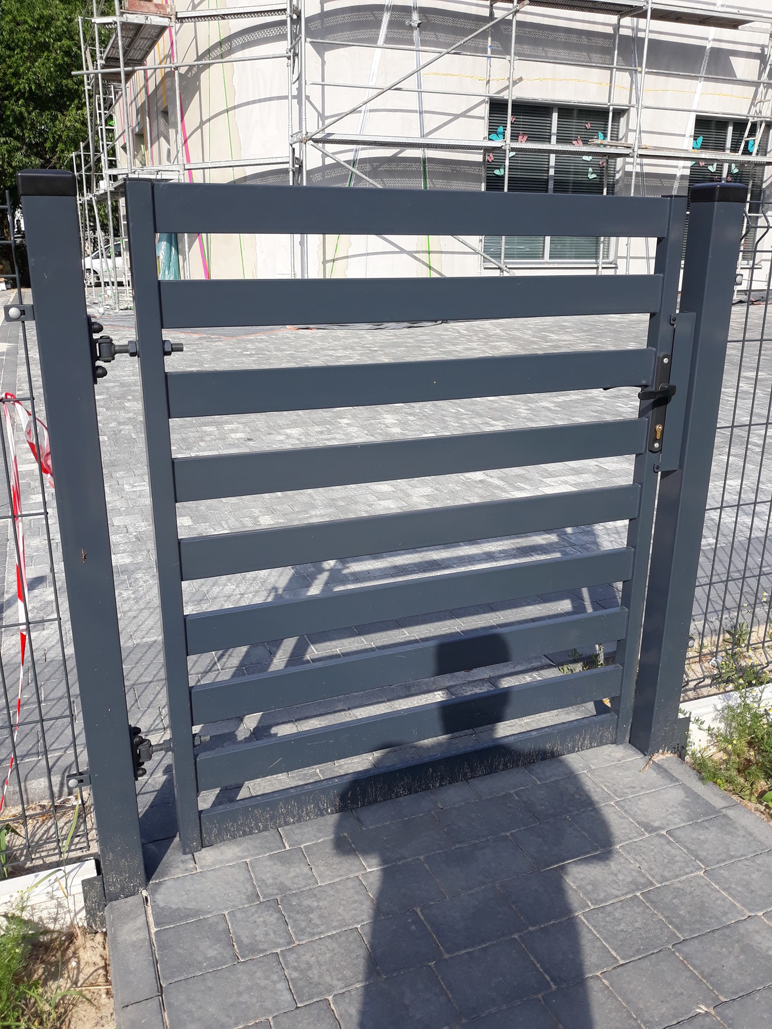 Grafitowa furtka z poziomych paneli, widoczna na tle placu budowy z rusztowaniami i dekoracjami okiennymi w kształcie motyli.