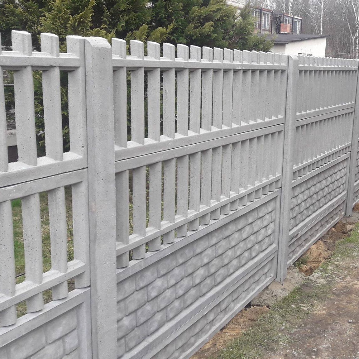 Szary betonowy płot z imitacją cegły w dolnej części i pionowymi szczebelkami w górnej, widoczne drzewa w tle.