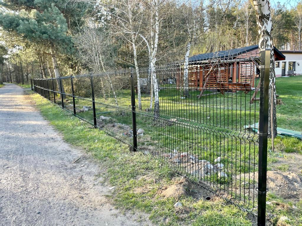 Nowoczesne, czarne ogrodzenie panelowe 3D, widok z boku wzdłuż ścieżki, z widocznym domkiem letniskowym w tle.