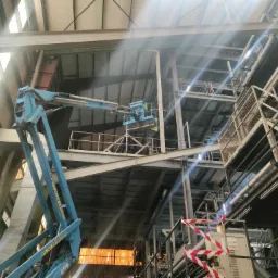 Wnętrze hali przemysłowej z widoczną konstrukcją stalową i podnośnikiem koszowym w trakcie prac na wysokościach, z pracownikami w żółtych kamizelkach w koszu, oświetlone promieniami słońca...