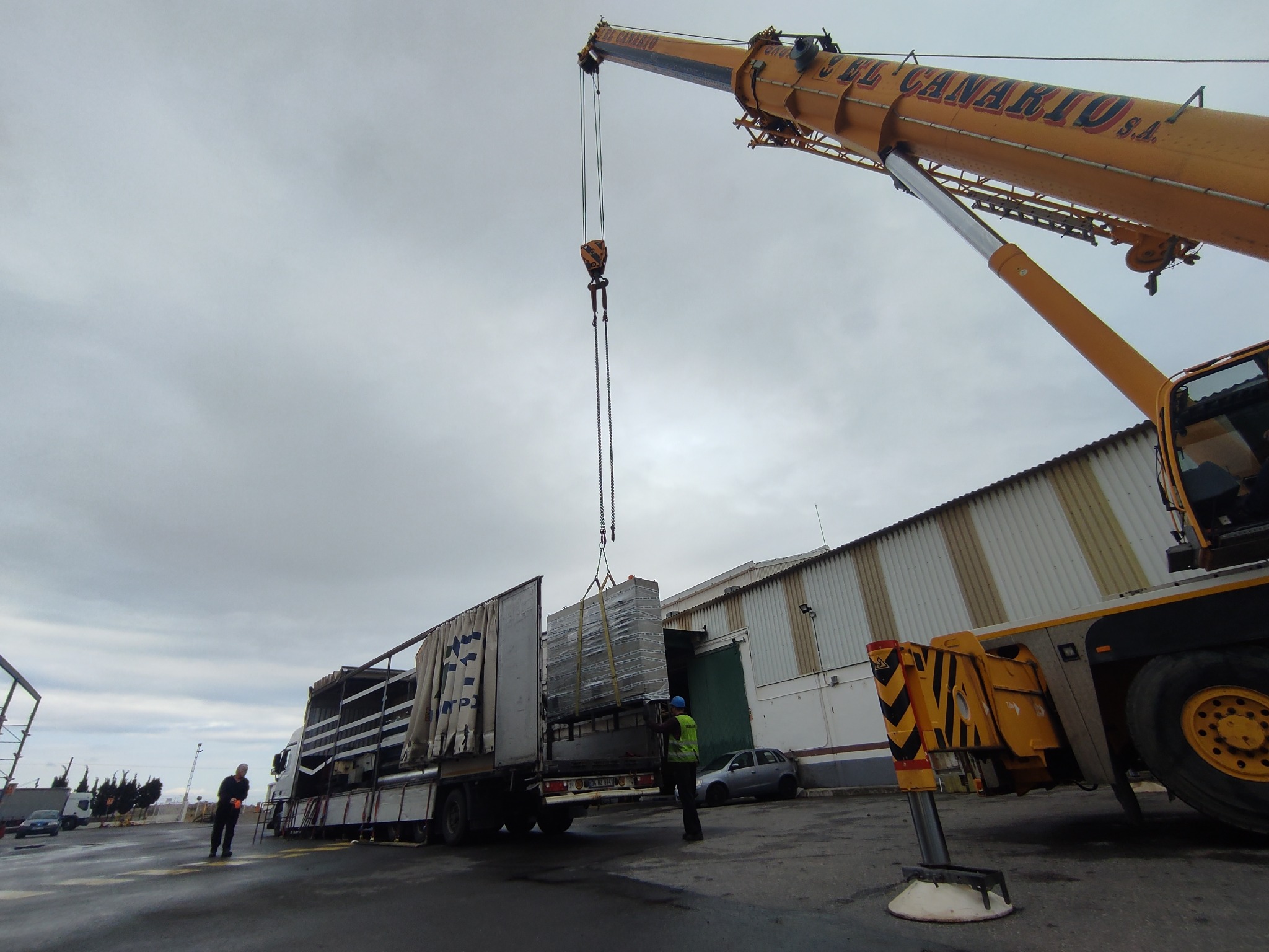 Żółty dźwig firmy El Canario S.A. podnosi pakiet materiałów budowlanych z ciężarówki, pracownik w kamizelce odblaskowej nadzoruje operację na tle hali magazynowej i pochmurnego nieba.