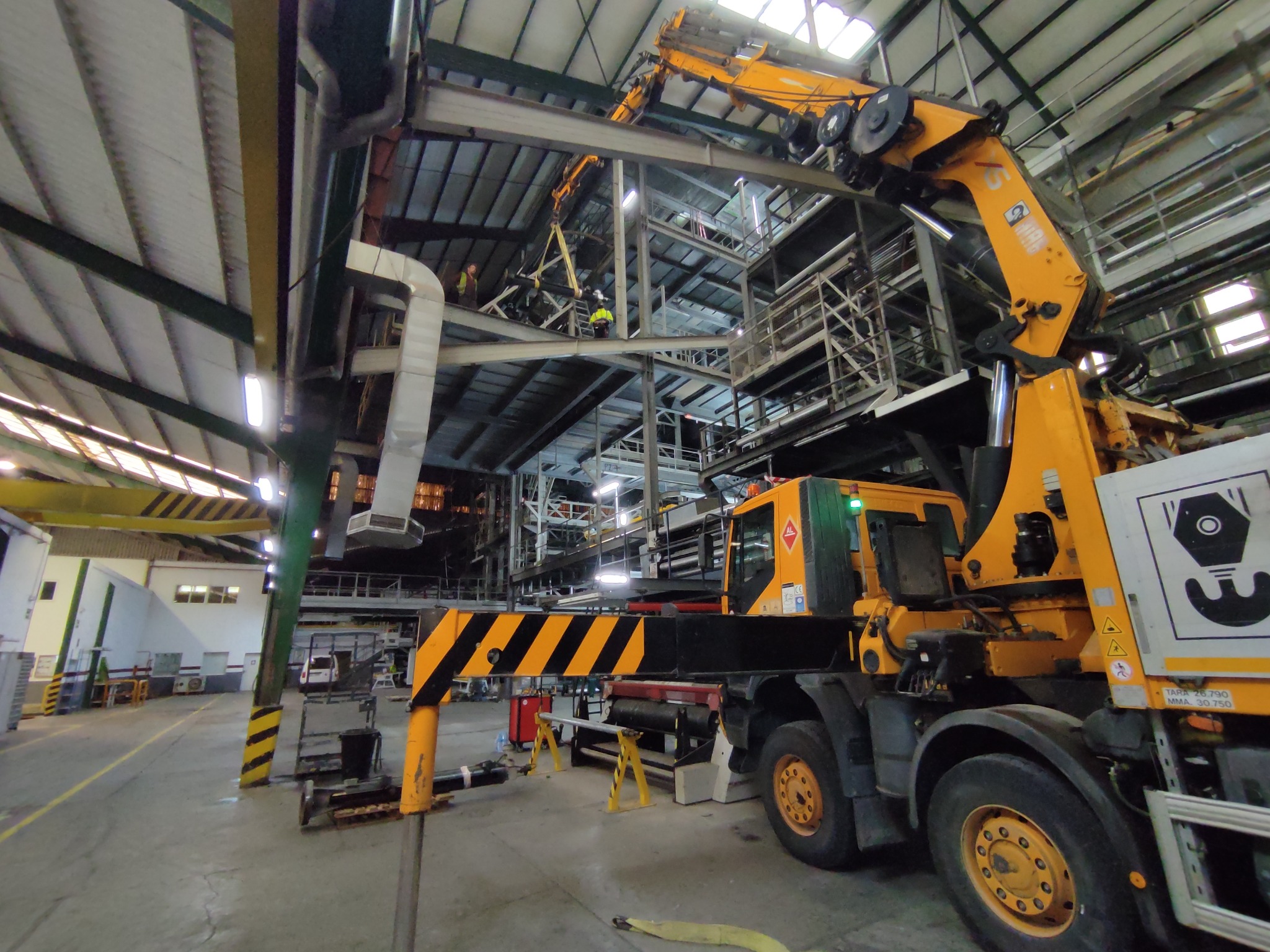 Żuraw samojezdny w hali przemysłowej podczas demontażu konstrukcji stalowej, pracownicy w odblaskowych kamizelkach na wysokości.
