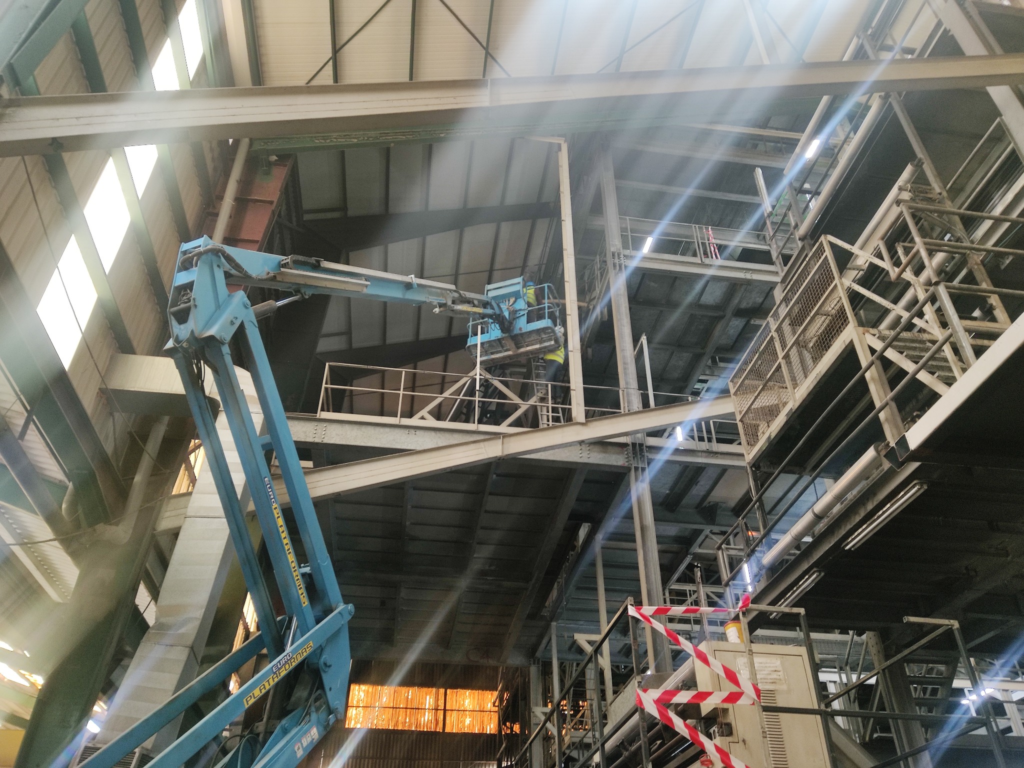 Wnętrze hali przemysłowej z widoczną konstrukcją stalową i podnośnikiem koszowym w trakcie prac na wysokościach, z pracownikami w żółtych kamizelkach w koszu, oświetlone promieniami słońca...