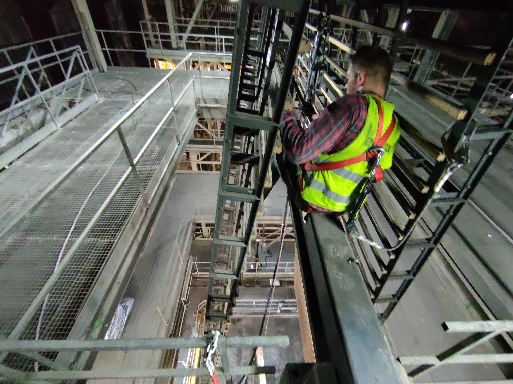 Pracownik w kamizelce odblaskowej, zabezpieczony linami, na metalowej konstrukcji wewnątrz budynku przemysłowego, widok z dołu na skomplikowany układ belek i platform.