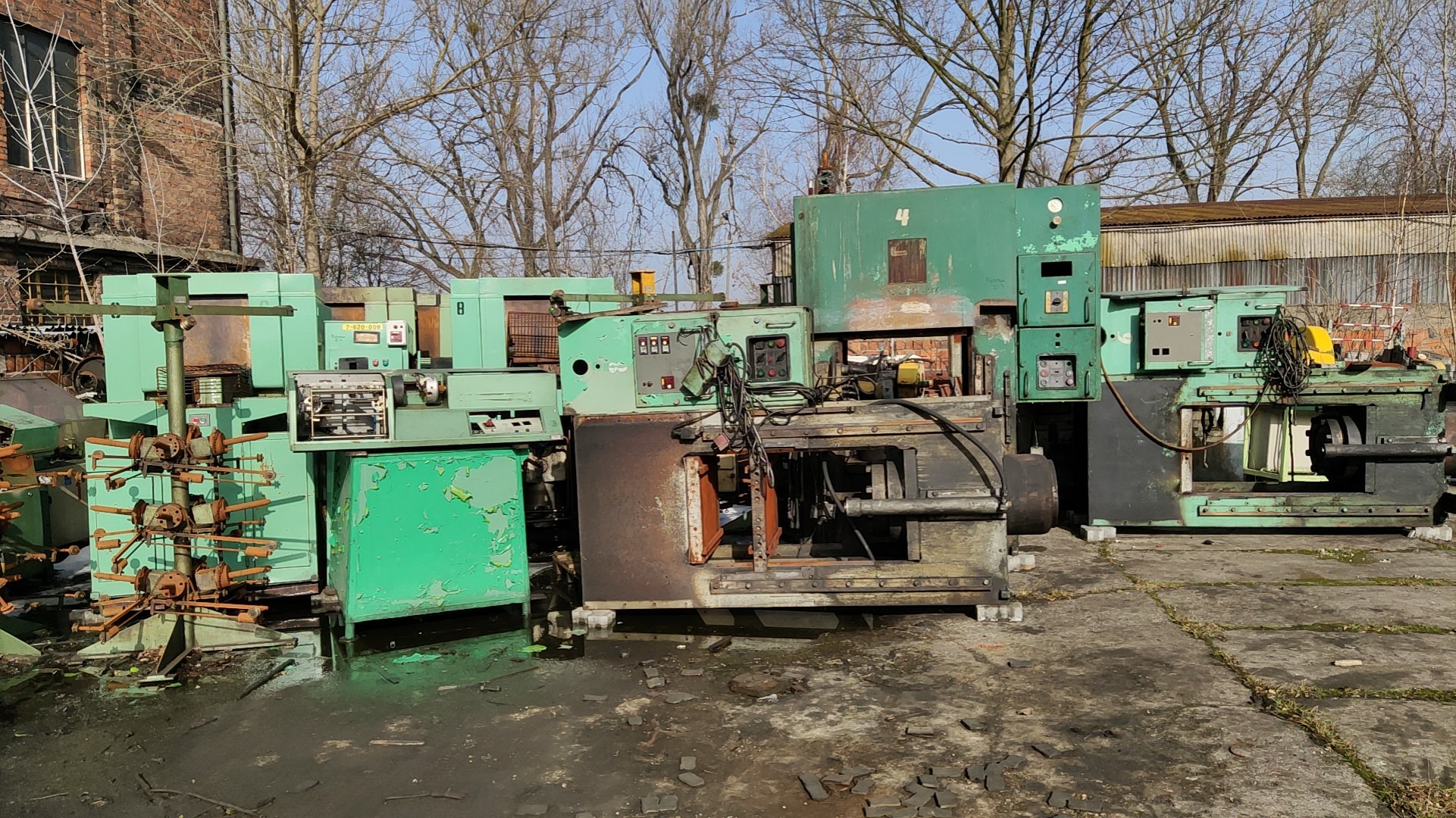 Zdemontowane, zielone maszyny przemysłowe, ustawione na zewnątrz w pobliżu ceglanego budynku i drzew bez liści.