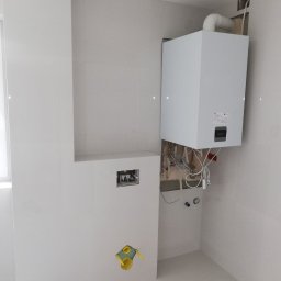 Wykończenia Marcin Kieryk Działalność usługowa - Biała łazienka w trakcie prac wykończeniowych: zamontowany piec gazowy z widocznymi rurami i przewodami, przygotowane miejsce na podtynkowy stelaż WC oraz instalacja wodno-kanalizacyjna.