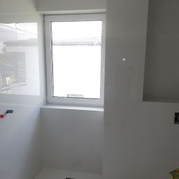 Wykończenia Marcin Kieryk Działalność usługowa - Wnętrze łazienki w trakcie prac wykończeniowych, widoczne białe płytki na ścianach i podłodze, okno z widokiem na zewnątrz oraz otwór odpływowy w podłodze zabezpieczony niebieską folią.