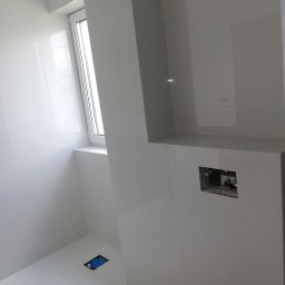 Wykończenia Marcin Kieryk Działalność usługowa - Białe, błyszczące płytki w łazience, widoczne otwory na instalacje, fragment okna z parapetem.