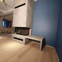 Wykończenia Marcin Kieryk Działalność usługowa - Nowoczesny kominek obłożony szarymi płytami, z czarnym wkładem kominkowym i przylegającą ławką, na tle pomalowanej na granatowo ściany i parkietu w jasnym odcieniu drewna.