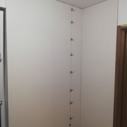 Wykończenia Marcin Kieryk Działalność usługowa - Biała ściana z zamontowanymi płytami gipsowo-kartonowymi, pionowy rząd metalowych klipsów utrzymujących płyty na miejscu, widoczne łączenia płyt, podłoga z paneli.