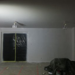 Wykończenia Marcin Kieryk Działalność usługowa - Pokój w trakcie malowania: ściany pomalowane na szaro, podłoga zabezpieczona folią, drzwi osłonięte czarną folią malarską, widoczne puszki z farbą i prowizoryczne oświetlenie.