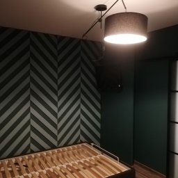 Wykończenia Marcin Kieryk Działalność usługowa - Pokój z dekoracyjną ścianą w jodełkę w odcieniach szarości i zieleni, widoczny stelaż łóżka bez materaca, oświetlony lampą sufitową z abażurem w kolorze szarym.