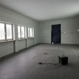 Duże, puste pomieszczenie wyłożone szarymi płytkami podłogowymi i białymi płytkami ściennymi, z trzema oknami po lewej stronie, zakrytymi parapetami, zasłoniętymi drzwiami z tyłu i grzejnikiem...